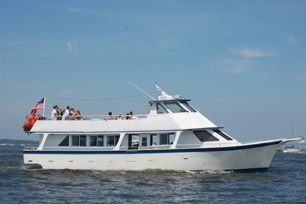 White passenger boat with people on the upper deck, sailing on a calm body of water under a clear blue sky. An American flag is displayed at the rear.